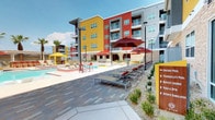 This expansive amenity space is defined by its bold, resort-inspired design, centered around a generously sized swimming pool with crystal-clear turquoise water. The pool area is framed by the striking multi-story building facade, clad in vivid red, yellow, and gray panels accented by stacked stone, creating a lively and energetic atmosphere.

The sun deck surrounding the pool is thoughtfully appointed with rows of modern lounge chairs fitted with cushioned sling seats, complemented by large red and yellow market umbrellas providing ample shade. Multiple seating zones invite relaxation, including a curved built-in bench area shaded by bold red shade sails, ideal for gathering. A dedicated outdoor kitchen station, finished in stacked stone with stainless steel grills and prep surfaces, sits beneath a white pergola, offering a well-equipped space for outdoor entertaining.

A separate fire pit lounge area features a built-in sectional sofa beside a linear blue glass fire feature, adding an intimate evening ambiance. Lush ornamental plantings and patterned tile pavers throughout the courtyards enhance the overall aesthetic.

Indoors, a game room features bold red columns, tile flooring, ping-pong and billiards tables, wall-mounted screens, and ceiling fans, providing a vibrant social gathering space.