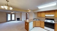 This single-floor residence offers a thoughtfully designed open-concept layout that seamlessly connects the kitchen and living areas, creating a spacious and cohesive environment. The kitchen is outfitted with warm honey-toned wood cabinetry, dark granite-style countertops, and an L-shaped peninsula that provides both ample prep space and a natural divider between the kitchen and living room. A built-in dishwasher and double sink add to the kitchen's functional appeal, while a skylight overhead floods the space with natural light. The living area features plush wall-to-wall carpeting and a fireplace that serves as a warm focal point, complemented by a ceiling fan and access to a glass-paned exterior door that invites natural light into the room. A textured ceiling treatment adds subtle visual interest throughout. A hallway leads to additional rooms beyond, hinting at a well-organized floor plan. The overall ambiance is comfortable and inviting, with neutral tones, warm wood accents, and a practical design that balances open living with defined functional zones.