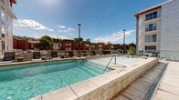 Located at 580 N 300 W in Salt Lake City, Utah, this outdoor amenity space serves as a vibrant recreational hub for the surrounding residential community. The centerpiece of the area is a generously sized, crystal-clear swimming pool framed by smooth stone coping and wide tiled decking, providing ample space for both relaxation and activity. Integrated directly alongside the pool is a dedicated hot tub or spa zone, complete with water jets, offering a contrast to the main pool. Surrounding the pool deck is a generous arrangement of cushioned lounge chairs and reclining sun loungers, ideal for enjoying Salt Lake City's abundant sunshine. Glass panel barriers and low perimeter walls define the space while maintaining open sightlines to the surrounding neighborhood and sky. An adjacent interior fitness or amenity room is accessible via large sliding glass doors, seamlessly connecting indoor and outdoor spaces. The modern multi-story residential buildings that frame the courtyard feature a bold contemporary design with mixed materials including white paneling, dark metal cladding, and red accent trim. The entire outdoor deck is finished in light-toned stone pavers, lending a clean, upscale aesthetic. The space enjoys expansive open-sky views and a bright, welcoming atmosphere year-round.