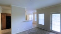 This single-floor residence offers a clean, open-concept layout designed for comfortable everyday living. The living area is carpeted and bathed in natural light, courtesy of multiple windows dressed with classic horizontal blinds that allow for adjustable brightness throughout the day. Crown molding runs along the ceiling, adding a refined architectural detail that elevates the overall aesthetic of the space.

A defining feature of the layout is the half-wall pass-through that connects the kitchen to the living area, creating a sense of openness while maintaining a subtle separation between spaces. The kitchen benefits from track lighting on the ceiling, providing focused illumination for food preparation and entertaining. The pass-through counter also offers an opportunity for casual seating or serving.

The neutral color palette throughout — from the warm beige walls to the light carpet — creates a calm and cohesive ambiance that is easy to personalize. The white trim and baseboards provide crisp contrast against the walls, contributing to the home's tidy, well-maintained appearance. The space receives generous natural light from multiple exterior-facing windows, lending a bright and airy feel. Overall, this residence combines thoughtful architectural details with a functional, inviting layout.