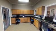 This single-floor business center is designed to support a productive and organized shared workspace environment. The room is finished with warm gray walls, crown molding, and a textured ceiling that adds a subtle architectural detail throughout. Underfoot, neutral wall-to-wall carpeting provides a comfortable and professional atmosphere.

The defining feature of this space is the extensive built-in L-shaped workstation that wraps around multiple walls, offering generous desk surface area topped with dark granite-style countertops. Rich, warm-toned oak cabinetry lines the walls both above and below the countertops, providing substantial storage for office supplies and equipment. Multiple computer workstations are integrated into the built-in desk, each accompanied by ergonomic task chairs with blue upholstery.

A printer and scanner are positioned along the counter, making the space fully equipped for day-to-day administrative tasks. A window fitted with horizontal blinds and a decorative valance allows natural light to filter in while maintaining privacy. An additional window with a pass-through view into an adjacent area adds connectivity to the broader facility.

Access is provided through a glass-paned interior door, contributing to an open and professional feel. Overhead fluorescent lighting ensures the workspace is consistently and evenly illuminated. Overall, the space functions as a well-appointed, self-contained business center suited for multiple simultaneous users.