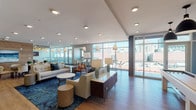 The 5th Floor Lounge is a thoughtfully designed communal amenity space that seamlessly blends comfort, functionality, and modern aesthetics. Floor-to-ceiling windows wrap around much of the space, flooding the interior with natural light and framing striking views of the surrounding urban skyline. Light hardwood flooring runs throughout, providing a warm, cohesive foundation that ties the various zones together.

The main lounge area features contemporary seating arrangements, including a neutral-toned sofa, accent chairs in leather and fabric, and layered blue area rugs that add depth and color. A wooden credenza anchors the space, complemented by decorative table lamps and curated accessories. Adjacent to the lounge, a dedicated dining area accommodates a large wooden conference-style table surrounded by upholstered chairs, set against a striking wood-accent wall with a mounted piece of artwork. A billiards table occupies its own nook nearby.

A casual high-top seating area with bistro tables offers additional gathering space beside the expansive windows. The lounge also includes a well-appointed kitchenette featuring white quartz countertops, dark cabinetry, a stainless steel refrigerator, and warm pendant lighting, making the space well-suited for both relaxed socializing and private events.