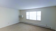 This single-floor space presents a clean, minimalist interior characterized by neutral wall tones and wall-to-wall carpeting in a warm beige hue. The main room is generously proportioned, offering an open and unobstructed layout suitable for a variety of furniture arrangements. A large, multi-pane window fitted with horizontal blinds dominates one wall, allowing an abundance of natural light to fill the room and contributing to its airy, open atmosphere. Climate control is provided by a wall-mounted air conditioning unit positioned adjacent to the window, complemented by a baseboard heating unit that runs along the exterior wall—ensuring year-round comfort. The walls are finished in a clean, off-white tone that enhances the brightness of the space. The overall design is straightforward and functional, with smooth ceilings and simple, unadorned surfaces that provide a neutral canvas for personalization. The combination of practical climate systems, ample natural light, and open floor space makes this a versatile and comfortable single-floor unit.
