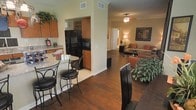 This single-story residence showcases an open-concept floor plan that seamlessly connects the kitchen, dining, and living areas, creating a cohesive and functional living environment. The rich, dark hardwood flooring runs throughout the main living spaces, providing a warm contrast to the light-toned walls adorned with crown molding details.

The kitchen is outfitted with medium-toned wood cabinetry extending to the upper walls, granite-look countertops, and black appliances including a side-by-side refrigerator and built-in microwave. A partial wall divides the kitchen from the living area while maintaining an open feel. A bistro-style breakfast bar with black metal bar stools offers casual dining directly adjacent to the kitchen.

The living room benefits from a ceiling fan with lighting, and is furnished to highlight its generous proportions. Large indoor plants add a natural element to the space. A decorative framed mirror anchors one wall, while warm ambient lighting from floor lamps enhances the inviting atmosphere. A hallway visible from the living area suggests additional rooms beyond, completing this well-appointed single-floor residence.
