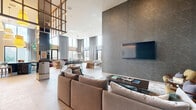 Located in San Diego, CA 92129, this elegantly designed clubroom delivers a sophisticated community gathering space with a generous open floor plan. Soaring double-height ceilings define the architectural character, creating an airy, voluminous atmosphere accentuated by floor-to-ceiling windows draped with floor-length curtains that flood the interior with natural light.

The space is thoughtfully zoned into distinct areas for socializing and recreation. A plush lounge area features a large sectional sofa arranged around a wall-mounted flat-screen TV set against a textured stone accent wall. A central gathering zone showcases woven accent chairs, a curved burgundy sofa, and a statement live indoor tree that adds organic warmth. A dedicated bar counter finished in rich dark marble with wood detailing serves as a striking focal point.

The game zone includes a billiards table framed by curated wall art and a foosball table, while a casual banquette with built-in bench seating and small café-style tables offers an ideal setting for informal meetings. Warm pendant lighting, terrazzo-style flooring, mixed-material wall treatments in muted greens and deep navy, and lush indoor greenery contribute to the refined yet welcoming ambiance throughout this dynamic clubroom.