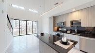 This contemporary two-story residence at 5211 Peachtree Blvd in Chamblee, Georgia, showcases a refined modern design throughout. The main level is anchored by a dramatic open-concept living and kitchen area defined by soaring double-height ceilings and strategically placed clerestory windows that flood the space with natural light. The kitchen is thoughtfully appointed with flat-front cabinetry in a warm light wood finish, dark granite countertops, stainless steel appliances including a gas range, built-in microwave, French door refrigerator, and dishwasher, all complemented by a dark tile backsplash. A generously sized central island with a matching dark granite surface provides additional prep space and seating. Light wood-tone flooring runs throughout, lending warmth and cohesion to the open layout. Full-height glass doors on the main level open to an outdoor area, enhancing the connection between interior and exterior. The upper level features a bright, open loft space with recessed lighting, a ceiling fan, and large windows offering views of the surrounding neighborhood. A modern cable railing system elegantly defines the loft's open edge overlooking the main floor below. The bathroom is finished with clean white tile, a glass-enclosed walk-in shower with a rain showerhead, and minimalist fixtures, reflecting the home's consistently sleek and contemporary aesthetic.