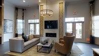 This single-floor space makes an immediate impression with its dramatic, soaring ceilings that lend an expansive, airy quality to the interior. The centerpiece of the lounge area is a sleek linear fireplace framed by striking floor-to-ceiling decorative panels composed of natural wood cross-sections, adding an organic, artisan touch to the otherwise contemporary design. A wall-mounted flat-screen television is seamlessly integrated into this focal wall, creating a cohesive and functional entertainment feature. Above the seating arrangement, a cylindrical chandelier with candelabra-style lighting anchors the space and adds a refined elegance. The lounge is furnished with a tufted gray sofa, a minimalist rectangular coffee table, and a pair of warm caramel-toned wingback chairs, striking a comfortable balance between style and function. Natural light pours in through multiple sets of glass-paneled doors and windows along the perimeter, connecting the interior to the outdoors while brightening the neutral gray walls. The flooring features a patterned area rug layered over hardwood, adding texture and warmth underfoot. Overall, the space exudes a polished yet inviting ambiance, thoughtfully designed for both relaxation and social gatherings.
