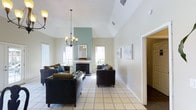 This single-story space centers around a spacious, open living area defined by soaring vaulted ceilings that create an airy, expansive atmosphere. A striking sage-green accent wall frames a classic white fireplace, serving as the room's focal point and adding a subtle pop of color against the otherwise neutral white walls. An elegant wrought-iron chandelier with warm-toned shades hangs prominently from the peak of the vaulted ceiling, complementing the overall design.

Natural light flows generously into the room through multiple windows on either side of the fireplace, brightening the light-toned ceramic tile flooring that runs throughout the main living space. Dark leather seating is arranged to create a comfortable and functional conversation area around a central coffee table.

Framed artwork adorns the walls, adding tasteful decorative touches, while a doorway on the right side of the living area leads to an adjacent room with contrasting darker flooring, suggesting additional living or functional space. The overall ambiance is clean, bright, and welcoming, combining practical design with comfortable, casual elegance.