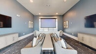 This well-appointed media lounge is designed to deliver an immersive entertainment experience within a refined, contemporary setting. The room is finished in a soothing blue-gray palette that extends across all walls, complemented by crisp white baseboards and a neutral ceiling with recessed lighting that ensures even, ambient illumination throughout. A ceiling-mounted projector anchors the primary viewing experience, paired with a large motorized projection screen positioned at the front of the room. A large flat-screen television mounted on the adjacent side wall provides an additional display option, supported by a sleek, light-toned media credenza beneath it. Seating is arranged in a theater-inspired configuration, with plush gray sofas and accent chairs flanking a long, low central table — ideal for group viewings or social gatherings. Patterned, dark-toned carpeting adds texture and visual depth underfoot, tying together the layered design. Gold-framed artwork adorns the walls, lending warmth and a curated aesthetic to the space. Large windows along the rear wall allow natural light to filter in and offer views of an outdoor patio area beyond, creating a seamless connection to the exterior. The overall ambiance is sophisticated yet inviting, balancing functionality with modern design sensibility.