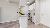 This well-designed single-floor space offers a clean, contemporary aesthetic throughout, characterized by light neutral tones, white trim, and consistent light wood-look flooring that unifies each area seamlessly.

The kitchen is a functional and stylish centerpiece, featuring crisp white shaker-style cabinetry, light stone countertops, and a convenient breakfast bar peninsula that opens toward the living area — ideal for both everyday use and casual entertaining. A gas range and stainless steel dishwasher complete the thoughtfully equipped cooking space.

The main living area is open and airy, benefiting from a large window dressed with vertical blinds that allows ample natural light to fill the room. A ceiling fan adds comfort, while exposed ceiling beams lend a subtle architectural detail to the otherwise clean-lined space.

The hallway connects the living and kitchen areas to the rest of the unit, lined with built-in white cabinetry offering generous storage. An electrical panel is conveniently recessed into the wall nearby.

The bathroom is neatly finished with the same light flooring, featuring a framed glass walk-in shower enclosure with a white surround and a standard toilet. Colorful towels add a welcoming touch to the otherwise serene, minimal palette. Overall, the space presents a bright, move-in-ready environment with cohesive modern finishes throughout.