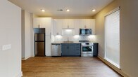 This single-floor unit in Durham, North Carolina, presents a clean, contemporary design with a cohesive aesthetic carried throughout the space. Wood-look plank flooring runs continuously from room to room, providing a warm, unified foundation that complements the crisp white walls and modern trim work.

The kitchen is a standout feature, designed with both style and functionality in mind. Two-toned cabinetry pairs white upper cabinets with deep slate-blue lower cabinets, accented by a classic white subway tile backsplash. Stainless steel appliances — including a refrigerator, range, microwave, and dishwasher — complete the space, which also benefits from natural light via a nearby window.

A central hallway connects the various rooms, lined with gray doors fitted with matte black hardware. The corridor leads to a well-lit living area featuring neutral warm-toned walls and a large window with blinds, allowing ample natural light to fill the room.

The bathroom maintains the modern theme, with matte black fixtures including a towel bar and toilet paper holder, alongside a built-in shelving unit that provides practical storage. An electrical panel is conveniently accessible along the hallway. The overall ambiance is clean, minimal, and move-in ready.