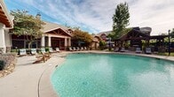 Located at Legacy Oaks Place in Asheville, North Carolina, this expansive outdoor amenity complex offers a resort-style experience set against a backdrop of mature hardwood trees and Blue Ridge Mountain surroundings. The centerpiece of the space is a large, freeform swimming pool with smooth curved edges, crystal-clear turquoise water, and multiple entry points, creating an inviting atmosphere for relaxation and recreation.

Surrounding the pool, a generous concrete deck provides ample room for teak-framed lounge chairs and seating areas, many accented with teal cushions that complement the water's color. Teal market umbrellas are positioned throughout for shade and comfort. The deck transitions seamlessly between the pool and the various architectural structures that frame the space.

The main clubhouse building features a craftsman-style design with stone and wood exterior accents, large column-supported overhangs, and floor-to-ceiling windows that allow natural light to flow through the interior. A separate open-air pavilion with exposed timber framing and a gabled roof provides shaded gathering space, complemented by a stone-accented hot tub or spa nearby. Lush, mature trees surround the entire area, offering privacy and a natural canopy that enhances the serene, park-like ambiance of this well-appointed community amenity space.