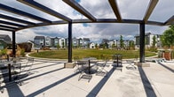 Located at 1901 S Hover Street in Longmont, Colorado, this community park serves as a central outdoor gathering space within a modern residential neighborhood. The park is anchored by a striking steel pergola structure with bold dark beams, providing partial shade over a spacious concrete patio furnished with bistro-style tables and chairs — an ideal setting for casual outdoor dining and socializing. Beneath the pergola, a built-in outdoor kitchen features multiple stainless steel grills set into a tiled base, offering a fully equipped space for community cookouts and gatherings. Adjacent to the covered patio, an expansive open lawn provides generous green space for recreation and relaxation under Colorado's wide open skies. A nature-inspired playground area incorporates wood chip surfacing, sculptural log climbing features, and a rock wall, creating an engaging environment for children. Winding concrete walkways meander throughout the park, lined with thoughtfully curated landscaping including ornamental grasses, native plantings, mulched beds, and young trees that will mature to provide additional shade and greenery. The surrounding contemporary multi-story residences frame the park on all sides, reinforcing its role as a vibrant communal hub within the neighborhood.