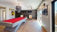 Located at 504 E Ninth St in Tucson, Arizona, this single-floor community space offers a thoughtfully designed interior that blends comfort, functionality, and eclectic style. The open living area is anchored by a large sectional sofa, recessed lighting, and a ceiling fan, with expansive windows that flood the room with natural light and offer views of the surrounding urban landscape. A sliding glass door connects the living space to an outdoor balcony, enhancing the sense of openness. The game room area is a standout feature, centered around a vivid red felt pool table beneath a dramatic multi-tiered chandelier. A bold black accent wall, wall-mounted televisions, classic upholstered armchairs, and additional sliding doors to the balcony give this zone a lively yet sophisticated atmosphere. The kitchen is equally impressive, showcasing rich dark wood cabinetry, granite countertops, and a striking chevron-patterned wood herringbone backsplash that serves as a bold design centerpiece. Stainless steel appliances, a commercial-grade coffee station, and ample counter space make it highly functional. Warm hardwood flooring runs consistently throughout, tying all areas together into a cohesive, inviting environment well-suited for gathering and socializing.