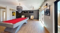 Located at 504 E Ninth St in Tucson, Arizona, this single-floor community space offers a thoughtfully designed interior that blends comfort, functionality, and eclectic style. The open living area is anchored by a large sectional sofa, recessed lighting, and a ceiling fan, with expansive windows that flood the room with natural light and offer views of the surrounding urban landscape. A sliding glass door connects the living space to an outdoor balcony, enhancing the sense of openness. The game room area is a standout feature, centered around a vivid red felt pool table beneath a dramatic multi-tiered chandelier. A bold black accent wall, wall-mounted televisions, classic upholstered armchairs, and additional sliding doors to the balcony give this zone a lively yet sophisticated atmosphere. The kitchen is equally impressive, showcasing rich dark wood cabinetry, granite countertops, and a striking chevron-patterned wood herringbone backsplash that serves as a bold design centerpiece. Stainless steel appliances, a commercial-grade coffee station, and ample counter space make it highly functional. Warm hardwood flooring runs consistently throughout, tying all areas together into a cohesive, inviting environment well-suited for gathering and socializing.