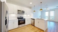 This thoughtfully laid-out one-bedroom, one-bathroom residence offers a clean, modern aesthetic throughout its single floor. The open-concept kitchen and living area serves as the heart of the home, anchored by a sleek white peninsula with an undermount sink and elegant gold pendant lighting overhead. The kitchen is appointed with crisp white shaker cabinetry, white subway tile backsplash, light quartz countertops, and a full suite of stainless steel appliances, including a French door refrigerator, electric range, over-the-range microwave, and dishwasher. Light wood-tone flooring runs throughout the main living areas, enhancing the bright, airy feel. A glass-paneled exterior door with sidelights welcomes natural light into the living space and offers views of the outdoors. The bedroom is carpeted for comfort and features a ceiling fan, a window with outdoor views, and access to a spacious walk-in closet with multi-level hanging rods. The bathroom is generously sized with wood-look flooring, a white vanity with ample drawer storage, a large mirror, and a soaking tub with shiplap-style tile surround. A window in the bathroom adds natural light to this clean, well-finished space.