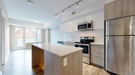 Located at 1140 Wellington St W in Ottawa's vibrant Hintonburg neighbourhood, this modern 1-bedroom, 2-bathroom unit offers a well-designed single-level layout with contemporary finishes throughout. The open-concept living and kitchen area serves as the heart of the unit, featuring a central island with a wood-toned base and clean quartz-style countertop, white upper cabinetry with subway tile backsplash, and a full suite of stainless steel appliances including a refrigerator, electric range, built-in microwave, and dishwasher. Track lighting illuminates the space, while large windows invite natural light into the living area. Light-toned luxury vinyl plank flooring runs consistently through the main living spaces and hallways, lending a cohesive, modern feel. The hallways are clean and well-proportioned, connecting the living area to the private bedroom and bathrooms. The bedroom is a comfortable, quiet retreat with a window offering natural light and ample wall space. The two bathrooms are finished with white ceramic tile; one features a soaker tub with tiled surround, while the other offers a glass-enclosed walk-in shower. Neutral grey tones throughout the unit create a calm, contemporary ambiance well-suited to a range of décor styles.