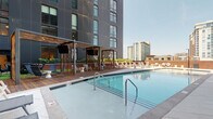 This outdoor amenity space is thoughtfully designed to deliver a resort-like experience within an urban high-rise setting. At its centerpiece sits a generously sized swimming pool with crisp, clear water framed by smooth tile coping and stainless steel entry rails. Along the pool's perimeter, a long row of sleek gray sling-style lounge chairs provides ample space for sunbathing, complemented by accent lighting for evening use.

Adjacent to the pool, a warm-toned hardwood deck serves as the foundation for a series of architecturally distinctive cabanas. Constructed with an open lattice framework of natural wood in a geometric cross-hatch pattern, the cabanas offer semi-private retreats equipped with comfortable lounge seating, side tables, and mounted screens for outdoor entertainment.

A separate courtyard zone extends the outdoor experience further, featuring large-format stone pavers, Corten steel raised planters filled with ornamental grasses, and intimate seating arrangements. Floor-to-ceiling glass walls allow visual connectivity to interior amenity spaces, including what appears to be a fitness center. Lush greenery, including manicured turf patches and climbing plants, softens the modern architecture throughout, while open sightlines to the surrounding cityscape reinforce the elevated urban setting.
