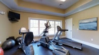 The fitness room at Mill is a dedicated exercise space designed to support an active lifestyle. The room is outfitted with multiple pieces of modern cardio equipment, including treadmills, an elliptical trainer, and a stationary bike, all arranged to allow comfortable movement between machines. A large multi-pane window spans much of one wall, flooding the space with natural light and offering views of the surrounding greenery, creating an inviting atmosphere for workouts. The flooring features durable rubberized material suited for fitness use, providing both traction and cushioning underfoot. Warm yellow-toned walls contribute to an energizing environment, while white trim and crown molding add a polished, finished look to the room. The ceiling incorporates recessed ventilation and lighting fixtures, ensuring the space remains comfortable during exercise. A white panel door provides private access, contributing to the room's clean, uncluttered aesthetic. The overall design balances functionality with thoughtful interior detailing, making this fitness space both practical and pleasant to use on a daily basis.