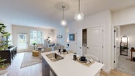 This single-floor residence offers a thoughtfully designed open-concept layout that seamlessly connects the kitchen, dining, and living areas. The kitchen is well-appointed with two-tone cabinetry—dark lower cabinets paired with white upper cabinets—complemented by light quartz countertops, a stainless steel appliance suite including a French door refrigerator, range, and microwave, plus light wood-style flooring throughout. A kitchen island with a double sink serves as a natural gathering point, illuminated by two globe pendant lights.

The living room features light hardwood flooring, large windows that invite generous natural light, and a glass-paneled exterior door offering outdoor views. The space conveys a warm, airy ambiance with neutral tones throughout.

The unit includes multiple bedrooms, each carpeted for comfort. One bedroom features a window with exterior views and ample dresser space, while another offers an en-suite layout. A dedicated office space with carpet and neutral walls provides a quiet, functional work area. An in-unit washer and dryer is conveniently accessible via a closet. The bathroom features a white vanity with quartz countertop, wood-look flooring, and clean, minimalist finishes, contributing to the overall modern and cohesive aesthetic of the residence.