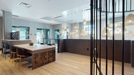 Located at 1 Midtown Bridge Approach in Hackensack, NJ, this single-floor amenity space showcases a sophisticated contemporary design that blends natural materials with modern architectural elements. The main lobby area immediately draws attention with its generous ceiling height, light hardwood flooring, and a striking cluster of woven rattan pendant lights that cascade at varying heights, creating a warm, textural focal point. A custom reception desk with intricate geometric wood paneling anchors the space, while sleek black metal partition frames define zones without sacrificing visual openness.

A lounge area features rattan egg chairs and a dramatic spiral staircase encased in a cylindrical black metal balustrade, adding sculptural interest. Nearby, a communal bar-height table with upholstered swivel chairs is framed by a decorative geometric screen wall, providing a semi-private gathering spot. Exposed concrete columns add an industrial contrast to the refined finishes throughout.

A separate entertainment lounge offers a decidedly different character, with a dark tray ceiling, Edison string lights, textured brick accent walls, bold retro-inspired murals, vibrant plaid carpeting, and a large mounted display screen — creating an energetic, casual atmosphere ideal for social gatherings.