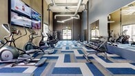 Located at 4722 E Bell Rd in Phoenix, AZ, this fitness center is a thoughtfully designed, single-floor facility offering two dedicated exercise rooms that together provide a comprehensive workout environment. The space features soaring ceilings with exposed industrial-style ductwork and large overhead fans, creating an open and energetic atmosphere.

The cardio and strength training room is finished with a bold blue and gray patterned carpet that adds a modern aesthetic. It is outfitted with treadmills, elliptical machines, and rowing equipment positioned near a large window overlooking an outdoor pool area, filling the room with natural light. A full dumbbell rack with an adjustable bench and wall-mounted mirrors complete this area, while multiple large flat-screen TVs keep users entertained throughout their workout.

The functional training room transitions to a vibrant green turf floor and features a heavy-duty multi-station rig with TRX suspension trainers, battle ropes, medicine ball racks, and resistance sled equipment. A large glass garage-style door floods the space with natural light and connects the interior visually to the outdoors. A Smith machine and additional wall-mounted mirrors round out the strength training offerings. A wall-mounted hand sanitizer station and WiFi access are also available, ensuring a convenient and clean workout experience.