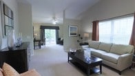 This single-floor residence showcases an open-concept living area defined by soaring vaulted ceilings that lend a spacious, airy quality to the interior. The living room is anchored by a sleek, dark-finished coffee table paired with a light-toned sofa, creating a balanced contrast that complements the neutral beige and gray color palette throughout. A ceiling fan with integrated lighting serves the main living area, providing both function and a subtle design element.

Large windows dressed with elegant valances and earth-toned draping panels allow generous natural light to fill the space while offering privacy. A sliding glass door in the background opens to what appears to be an outdoor patio or balcony, framing views of lush greenery and further enhancing the connection between indoor and outdoor living.

The adjoining sitting or dining area is visible beyond a partial wall, furnished with additional seating and a small table, suggesting a versatile layout suited for both relaxation and entertaining. Soft, wall-to-wall carpeting in a warm neutral shade runs throughout, contributing to the cohesive and comfortable ambiance of the home. Tasteful artwork and accent lighting complete the polished, move-in-ready aesthetic.