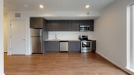 This single-floor unit offers a thoughtfully designed open-concept layout finished with warm hardwood-style flooring throughout. The kitchen is a standout feature, outfitted with sleek dark gray flat-front cabinetry, crisp white countertops, a textured tile backsplash, and a full suite of stainless steel appliances including a refrigerator, electric range, built-in microwave, and dishwasher. Recessed lighting above the kitchen provides bright, even illumination, complementing the clean, modern aesthetic. The living area is generously proportioned and bathed in natural light through large windows that frame views of the surrounding neighborhood. A wall-mounted HVAC unit ensures year-round climate control, and recessed ceiling lighting adds to the airy, well-lit atmosphere. The bathroom is finished in crisp white with a full soaking tub and shower combination, chrome fixtures, and coordinating hardwood-style flooring, offering a clean and functional space. A well-lit hallway connects the living spaces to the entryway, featuring recessed lighting, clean white walls, and ample room for smooth circulation. Throughout the unit, the consistent use of neutral tones, modern fixtures, and quality finishes creates a cohesive and contemporary living environment.