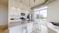 This contemporary single-floor residence showcases a clean, modern aesthetic throughout, anchored by light wood-tone flooring and a bright, neutral color palette. The open-concept kitchen is a standout feature, equipped with stainless steel appliances including a refrigerator, electric range, and microwave, all complemented by crisp white shaker cabinetry and sleek gray quartz countertops. A spacious island with bar seating serves as both a functional workspace and casual dining area. Exposed industrial-style ductwork and Edison-bulb pendant lights add an urban edge, while expansive floor-to-ceiling sliding glass doors flood the space with natural light and open to a private balcony offering sweeping city views. The two bedrooms are serene and well-proportioned, each with light flooring and neutral walls that create a calm, restful atmosphere. The bathrooms are thoughtfully designed — one features a glass-enclosed walk-in shower with built-in shelving for storage, while the second offers a generous white vanity with ample drawer space and a large mirror. A clean hallway connects the private quarters, and the overall layout flows efficiently, making excellent use of the available space throughout.