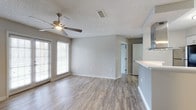 This single-floor space presents a well-designed open layout that combines comfort and functionality. The main living area is finished with wood-style laminate flooring in a warm gray tone, providing a clean and contemporary foundation throughout. Neutral gray walls create a versatile backdrop, complementing the bright, airy atmosphere enhanced by large windows fitted with horizontal blinds that allow natural light to filter in generously.

A ceiling fan is centrally positioned in the living area, offering both airflow and ambient lighting. The textured popcorn ceiling adds a classic touch overhead. Smooth white baseboards frame the flooring and contribute to the space's clean, finished appearance.

An open passageway connects the living area to additional rooms, suggesting a functional flow between spaces. A partial view of a white countertop to one side hints at a kitchen area nearby, while a door visible in the background leads to further rooms within the unit.

The overall ambiance is bright, neutral, and move-in ready, making the space adaptable to a variety of furnishing styles. The single-floor design ensures convenient, step-free living throughout.