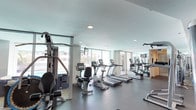 This fitness center offers a clean, contemporary workout environment designed to accommodate a variety of exercise needs. The space is characterized by its bright, open layout, enhanced by large floor-to-ceiling windows along the perimeter that flood the room with natural light and provide views of the surrounding exterior. The smooth, light-toned ceiling features recessed lighting, complementing the natural illumination throughout.

The cardio section is well-stocked with multiple treadmills and upright stationary bikes, all appearing to be modern Precor equipment. A multi-station cable and weight machine is positioned near the windows on one side, offering a comprehensive strength training option. A mirrored wall on one end visually expands the space and allows users to monitor their form during workouts. The flooring consists of durable, dark grey material suited for high-traffic fitness use.

The layout is thoughtfully organized, with adequate spacing between machines to ensure comfort and ease of movement. The overall ambiance is clean, minimalist, and functional, reflecting a professional fitness facility standard. A storage unit near the mirror wall provides additional utility to the space.