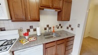 This single-floor residence offers a practical and well-organized layout, as seen in the overhead floor plan view. The kitchen is a standout feature, equipped with warm medium-toned wood cabinetry both above and below the counters, providing generous storage. Granite-look countertops complement the stainless steel sink, while a patterned tile backsplash featuring a cream and brown checkerboard accent detail adds visual interest. White appliances, including a dishwasher and range, complete the space.

The living area is generously sized, finished with neutral carpet and tile flooring at the entryway. A large window fitted with horizontal blinds allows natural light to filter in, and a wall-mounted air conditioning unit provides climate control. The entry door features warm wood tones consistent throughout the unit.

The bedroom is a comfortable, carpeted room equipped with a ceiling fan and adequate ventilation, maintaining the home's neutral color palette. The bathroom includes a single vanity with an integrated sink, a mirrored medicine cabinet offering additional storage, and chrome fixtures. A carpeted hallway connects the rooms, reinforcing the cohesive and functional flow of this single-level space.