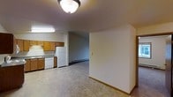 This single-floor residential space offers a practical and straightforward layout designed for comfortable everyday living. The kitchen is equipped with warm-toned wooden cabinetry, a white refrigerator, a dishwasher, and a countertop workspace, all set atop durable tile flooring that extends through the main living area. A fluorescent light fixture illuminates the kitchen zone, while a ceiling-mounted flush light provides additional brightness to the adjacent space. The open flow between the kitchen and the living area creates a connected, functional environment. The living area features carpet flooring and is warmed by a baseboard heating unit along the wall, making it well-suited for colder climates. Neutral-toned walls throughout the space lend a clean, understated aesthetic. A hallway or secondary room is visible through an open doorway, suggesting additional private spaces beyond the main area. The overall ambiance is practical and uncluttered, with a layout that maximizes usability. This space is well-suited for those seeking a no-frills, move-in-ready environment with essential amenities conveniently arranged.