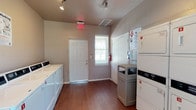This shared laundry facility is a practical, single-floor space designed to accommodate residents' laundering needs with efficiency and ease. The room features warm hardwood-style flooring throughout, providing a clean and durable surface well-suited for high-traffic use. A row of full-sized washing machines lines one wall, while stacked dryer units are positioned along the opposite side, maximizing the available floor space and offering ample capacity. A central payment kiosk is conveniently located near the dryers, enabling easy card-based transactions. A dedicated exterior door provides direct access to the facility, accompanied by a window fitted with blinds that allows natural light to filter into the space. The walls are finished in a neutral tone, contributing to a tidy and utilitarian atmosphere. Track lighting and ceiling-mounted fixtures ensure the room is well-lit at all times. Safety is addressed through the presence of a prominently displayed fire extinguisher and an illuminated emergency exit sign. A waste receptacle is centrally placed for resident convenience. Informational signage is posted near the machines, offering guidance on machine operation and payment instructions. Overall, this laundry facility is thoughtfully laid out to serve its functional purpose with clarity and comfort.