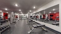 Located at 31 Walter Ave in Highland Park, NJ, this fitness center is a thoughtfully designed, single-floor facility built to accommodate a comprehensive workout experience. The space is defined by a bold red and white color scheme, with vibrant red accent walls contrasting cleanly against white surfaces and structural columns throughout.

The main workout floor is generously proportioned, finished with durable dark rubber flooring that provides traction and impact absorption across all exercise zones. Large wall-mounted mirrors span multiple sections of the room, visually expanding the space while allowing users to monitor their form during training sessions.

The strength training area is well-appointed with cable machines, multi-functional weight stations, adjustable benches, and a full rack of kettlebells and free weights. The cardio zone features multiple treadmills and an elliptical machine, all positioned beneath wall-mounted flat-screen televisions for entertainment during workouts. Stability balls and resistance accessories are also available for functional training.

Recessed overhead lighting ensures the entire floor is evenly and brightly illuminated. The facility includes separate entry and interior doors, with clearly marked emergency exits. The clean, organized layout offers ample open floor space between equipment stations, making the environment accessible and easy to navigate for users of all fitness levels.