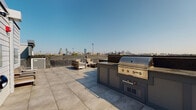 Located at 13 Shetland St in Boston's Roxbury neighborhood, this rooftop amenity space offers a well-appointed outdoor environment with striking views of the Boston skyline. The rooftop terrace is finished with large-format porcelain tile pavers that create a clean, contemporary aesthetic throughout the open-air area.

The outdoor kitchen is a standout feature, equipped with a built-in stainless steel grill and integrated cabinetry, making it an ideal setup for entertaining. Comfortable lounge seating is arranged nearby to take full advantage of the panoramic cityscape. The rooftop's low perimeter walls frame unobstructed sightlines across the Boston skyline, bathed in natural sunlight under open skies.

Access to the rooftop is provided through a modern glass-and-steel door system, transitioning from a clean, neutrally finished interior corridor. The interior access hallway features tile flooring consistent with the rooftop, neutral gray walls, and crisp white trim, maintaining a cohesive modern design language throughout. Safety features including fire suppression equipment are present near the rooftop access point.

Overall, this rooftop space combines functional outdoor amenities with a refined urban aesthetic, offering an inviting setting for relaxation and social gatherings with Boston's skyline as a backdrop.