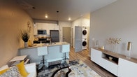This single-floor residence showcases a thoughtfully designed open-concept layout that seamlessly connects the living, dining, and kitchen areas. The kitchen is outfitted with stainless steel appliances, including a refrigerator, electric range, and built-in microwave, complemented by dark lower cabinetry, white upper cabinets, and a decorative tile backsplash. A peninsula with a built-in sink and seating for bar stools serves as both a functional workspace and casual dining area, illuminated by stylish pendant lighting overhead. Wood-style plank flooring extends throughout the main living area, adding warmth to the neutral gray and white color palette.

A stacked washer and dryer unit is conveniently situated within the unit, tucked neatly near the entry area. The bedroom features carpet flooring, a window with blinds for natural light, and ample wall space for storage and decor. The bathroom is finished with a dark wood vanity, a wide quartz-style countertop, an undermount sink, chrome fixtures, and a large frameless mirror with vanity lighting above. Clean-lined gray interior doors and recessed lighting throughout complete the modern, cohesive aesthetic of this well-appointed space.
