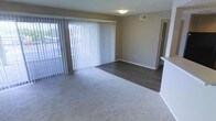 This single-floor space presents an open and functional layout designed for comfortable everyday living. The main living area is defined by a combination of plush carpeting and dark hardwood-style flooring, creating a clear visual distinction between zones within the room. Neutral beige-toned walls complement the flooring choices, contributing to a clean and versatile aesthetic throughout.

A large sliding glass door fitted with vertical blinds serves as a prominent feature of the living area, allowing natural light to filter into the space and providing direct access to an outdoor area. Glimpses through the door suggest an open exterior view, adding a sense of connection to the outdoors.

The textured ceiling adds subtle dimension overhead, while a centrally mounted flush light fixture provides practical overhead illumination. A wall-mounted HVAC vent ensures climate control convenience. White baseboards frame the perimeter of the room, offering a crisp contrast to the warm wall tones.

A partial wall or countertop edge visible on the right side hints at an adjacent kitchen or dining area, suggesting a functional flow between spaces. A doorway on the far wall leads further into the unit, indicating additional rooms beyond the main living area.