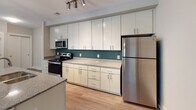 Located at 257 Broadway St in the heart of Asheville, NC, this single-floor unit offers a thoughtfully designed layout with modern finishes and a clean, contemporary aesthetic. The open-concept kitchen and living area serves as the centerpiece of the space, anchored by a granite-topped island with pendant lighting overhead and a ceiling fan that enhances airflow throughout. The kitchen is outfitted with floor-to-ceiling white cabinetry, granite countertops, a teal accent backsplash, and a full suite of stainless steel appliances, including a range, microwave, and refrigerator. Light wood-tone flooring runs throughout the main living areas, lending warmth and continuity. The bedrooms are carpeted for comfort and receive ample natural light through large windows with wooded views. One bedroom features a stylish barn-style sliding door, while another opens directly to a private balcony via sliding glass doors. The bathrooms are finished with granite-topped vanities, tile flooring in neutral tones, and accessibility-friendly grab bars. The overall ambiance is bright, modern, and functional, with thoughtful design details that balance style and practicality throughout the entire space.