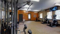 This single-floor fitness center offers a practical and energizing environment designed to support a full range of workout routines. The space is defined by its bold orange accent walls, which contrast with neutral beige tones to create a vibrant, motivating atmosphere. Patterned carpeted flooring in warm gold and beige tones covers the workout area, adding a comfortable underfoot surface throughout the room. Overhead, flush-mounted fluorescent light fixtures and a ceiling fan provide consistent illumination and airflow, keeping the space bright and well-ventilated. Natural light filters in through windows dressed with horizontal blinds, contributing to an open and airy feel. The room is furnished with a variety of fitness equipment, including a cable strength training machine, free weights, an upright stationary bike, and at least one elliptical or stair-climber machine, offering options for both cardio and resistance training. A framed piece of artwork decorates one wall, adding a touch of character to the functional space. Two doors — one solid wood and one with a window — provide access and visibility into adjacent areas. The overall layout is spacious enough to allow for comfortable movement between stations, making this fitness center a well-rounded facility for daily exercise.