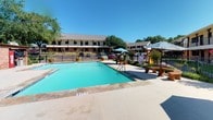 This Sherman property is centered around a generously sized outdoor swimming pool that serves as the focal point of the grounds. The pool reaches a depth of 8 feet and is surrounded by a broad concrete deck finished with stone tile edging, providing ample space for lounging. Multiple umbrella-shaded seating areas are positioned poolside, offering comfort during sunny Texas days.

The surrounding two-story brick building features covered walkway balconies along its exterior, framing the courtyard with a classic, structured appearance. A prominent wooden pergola anchors one side of the outdoor area, casting patterned shade across a paved patio beneath. Built-in bench seating and stone accent tables are arranged under and near the pergola, creating a relaxed gathering space.

A well-landscaped interior courtyard sits adjacent to the pool area, enclosed by wrought iron fencing. Lush plantings, flowering shrubs, trimmed hedges, and maintained green lawn fill the space, contributing to a welcoming and manicured outdoor environment. Mature trees throughout the property provide additional shade and a sense of established greenery. The overall ambiance is open, well-kept, and oriented toward outdoor leisure and communal enjoyment under the open Texas sky.