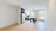 This thoughtfully designed single-floor unit offers a compact yet functional layout that maximizes every square foot. The open-plan living and kitchen area serves as the heart of the space, where contemporary design elements come together seamlessly. The kitchen is outfitted with a striking two-tone cabinetry scheme — crisp white upper cabinets contrast boldly with dark lower cabinets — complemented by a herringbone-patterned tile backsplash and a clean white countertop. Stainless steel appliances, including a refrigerator, range, microwave, and dishwasher, provide a full suite of modern conveniences. A linear pendant light fixture anchors the kitchen area, while recessed lighting adds ambient illumination. Large windows flood the living space with natural light and offer views of the surrounding urban streetscape and greenery. Light wood-tone flooring extends throughout the unit, lending warmth and visual continuity. The bedroom is a quiet, well-lit room featuring recessed ceiling lights, louvered closet doors, and clean white walls. The bathroom is finished in an all-white palette with a combination tub and shower, a sleek vanity with under-sink storage, white quartz countertop, and a modern rounded-rectangle mirror with integrated lighting.