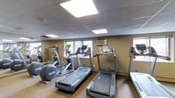 This fitness center offers a practical and functional workout environment designed to meet a range of exercise needs. The space is finished with a drop ceiling fitted with large recessed fluorescent light panels, providing bright, even illumination throughout. Warm-toned walls lend a neutral, focused atmosphere conducive to exercise.

The cardio equipment lineup is the centerpiece of the room, featuring multiple treadmills, an elliptical cross-trainer, a stationary upright bike, and what appears to be a recumbent bike or rowing machine along the far wall. The machines are thoughtfully arranged to allow comfortable use without overcrowding the space. A wall-mounted air conditioning unit ensures climate control during workouts.

Windows along one wall allow natural light to filter into the room, adding a sense of openness. The flooring features a low-pile carpet, providing a practical surface that absorbs sound and offers a degree of comfort underfoot. A wall-mounted mirror on one side visually expands the room while allowing users to monitor their form. Overall, this fitness center is a well-organized, amenity-ready space suited for cardiovascular training and general fitness maintenance.
