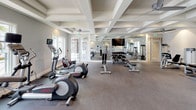 Located in Davenport, Florida, this single-floor fitness center offers a comprehensive and thoughtfully designed workout facility. The space is defined by its striking coffered ceilings throughout, which lend an architectural elegance uncommon in typical gym environments. Ceiling fans are integrated throughout, ensuring comfortable airflow during workouts.

The main exercise floor is carpeted and generously proportioned, housing an impressive array of cardio equipment including treadmills, elliptical machines, and a rowing machine. A wall of mirrors expands the sense of space, while a wall-mounted flat-screen TV provides entertainment. Ample natural light streams in through large windows, with French doors offering direct access to the outdoors.

A dedicated strength training area features a full complement of cable machines, weight benches, and a dumbbell rack, providing options for a complete resistance workout. Separate from the main floor, a bright cycling studio features light hardwood flooring, stationary bikes equipped with individual screens, and expansive windows that frame views of the surrounding landscape. This studio's open layout also makes it suitable for group fitness or yoga classes.

The facility's neutral gray palette, recessed lighting, and clean finishes create a bright, motivating atmosphere well-suited for a wide range of fitness activities.