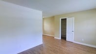 This single-floor space presents a straightforward and functional layout designed with practicality in mind. The interior is finished with warm hardwood flooring that runs throughout, adding a natural, inviting feel underfoot. The walls are painted in a soft, neutral palette — crisp white on one side transitioning to a warm golden yellow accent wall — creating a subtle contrast that brightens the overall atmosphere. The textured ceiling adds a classic touch consistent with the space's traditional character. A standout feature of the main room is the open built-in shelving unit, which offers generous storage and display potential without taking up additional floor space. Adjacent to the shelving, a white paneled door provides access to a separate room or closet area, contributing to the overall organizational functionality of the space. Clean white baseboards frame the flooring neatly throughout, lending a polished, finished look to the interior. The space is vacant and move-in ready, offering a blank canvas for a variety of furnishing styles and personal touches. Overall, the layout is efficient and well-maintained, making it a versatile option suited to a range of practical living arrangements.