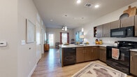 This well-designed one-bedroom, one-bathroom residence in San Antonio, TX offers an efficient and thoughtfully laid-out single-floor plan. The open-concept kitchen and living area is a standout feature, showcasing rich dark-walnut cabinetry, granite countertops, and a peninsula island with seating for casual dining. Sleek pendant lighting, recessed ceiling lights, and black appliances complete the modern kitchen aesthetic. Light-toned hardwood-style flooring flows throughout the main living areas, enhancing the sense of openness. The living room receives ample natural light through large windows dressed with bold accent curtains, and a ceiling fan adds comfort overhead. A door from the living area provides direct outdoor access. The spacious bedroom is carpeted for warmth and comfort, featuring a ceiling fan, natural light from a dedicated window, and ample room for furnishings. The full bathroom includes a tub-shower combination with marble-style tile surround and a vanity with granite countertops, all finished with wood-toned cabinetry consistent with the rest of the unit. A hallway and closet provide practical storage solutions. The overall ambiance is modern and inviting, with a neutral color palette throughout.
