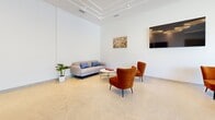 Located at 1370 Stittsville Main St in Ottawa, Ontario, this contemporary single-floor communal lounge offers a bright and thoughtfully designed gathering space. The open-plan layout is anchored by expansive floor-to-ceiling windows that flood the interior with natural light and provide sweeping views of the mature tree-lined streetscape beyond.

The living area is elegantly appointed with a mix of plush seating, including a textured grey sofa with blue accent cushions, vibrant burnt-orange armchairs, and sleek bistro-style dining tables with black chairs — creating distinct zones for relaxing and socializing. Light-toned large-format tile flooring runs throughout, enhancing the airy, clean aesthetic. A tray ceiling with recessed lighting adds architectural interest and a refined finish overhead.

Curated wall art, including a bold floral canvas, adds warmth and character to the crisp white walls. A wall-mounted television and decorative clock further contribute to the functional yet welcoming atmosphere. Toward the rear, a well-equipped kitchenette features warm wood cabinetry, built-in appliances, and a marble-effect backsplash. Slate-blue double doors provide access to additional amenities. The overall ambiance is modern, comfortable, and versatile — well-suited for both quiet relaxation and communal interaction.