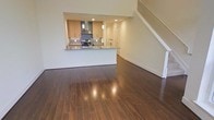 This contemporary two-story residence is designed with an open, flowing layout that maximizes both space and functionality. The main floor features rich, dark hardwood flooring that spans the living area, reflecting light and adding warmth to the interior. The open-concept design connects the living space seamlessly to the kitchen, which is outfitted with light wood cabinetry, granite or stone countertops, and stainless steel appliances including a gas range. A mounted range hood serves as a functional centerpiece above the cooktop, while pendant lighting adds a refined touch over the kitchen area. The kitchen counter extends into a breakfast bar, creating a natural divide between the cooking and living spaces while maintaining an airy, connected feel. Recessed lighting throughout the main floor ensures consistent, modern illumination. A staircase with clean white railings rises from the main floor to the upper level, where additional living quarters are situated. The neutral wall tones throughout complement the warm flooring and cabinetry, creating a cohesive and inviting ambiance. This well-appointed two-story residence offers a thoughtful balance of style and practicality, suited for comfortable everyday living.