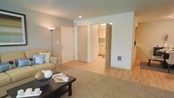 This single-floor residence offers a thoughtfully arranged living space finished in a calming palette of soft greens and warm neutrals. The living area serves as the central hub of the home, featuring a combination of plush carpeting and smooth hardwood-style flooring that delineates functional zones within the open layout. Recessed ceiling lighting and a tall floor lamp provide layered illumination throughout the space.

The living area comfortably accommodates a cushioned sofa accented with decorative pillows and a dark-toned coffee table, creating an inviting setting for relaxation. A wide entryway connects the living space to the kitchen area, which is visible in the background and features light wood cabinetry and ample counter space, suggesting a clean and functional cooking environment.

Two interior doors are positioned along the far wall, offering access to additional rooms or storage areas. A utility access panel is mounted low on the wall, indicating practical infrastructure within the unit. The overall design is clean and understated, with consistent trim work and white door panels throughout, contributing to a cohesive and well-maintained aesthetic.