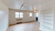 This single-floor space offers a clean, functional layout with an open and airy feel throughout. The main living area is bathed in natural light from multiple windows, highlighting the light-toned hardwood flooring that runs seamlessly across the space. A ceiling fan with integrated lighting occupies the center of the room, complemented by a track lighting system mounted near the entrance for added illumination and versatility. The walls are finished in a crisp, neutral white, creating a bright and welcoming atmosphere that makes the space feel open and adaptable. A curved, built-in shelving unit on one side adds a distinctive architectural touch, offering both display and storage potential. Baseboard heating ensures year-round comfort. Transitioning toward the back of the unit, an open passageway leads into a compact kitchen equipped with a stainless steel gas range and a sink, providing the essentials for everyday cooking. The textured ceiling adds subtle visual character throughout. Overall, the space presents a straightforward and practical layout, well-suited for those seeking a comfortable and low-maintenance living environment with ample natural light and thoughtful built-in details.